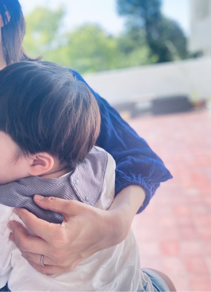 ベランダで子どもを抱きしめる母の腕。優しい光と初夏の緑が背景に映る