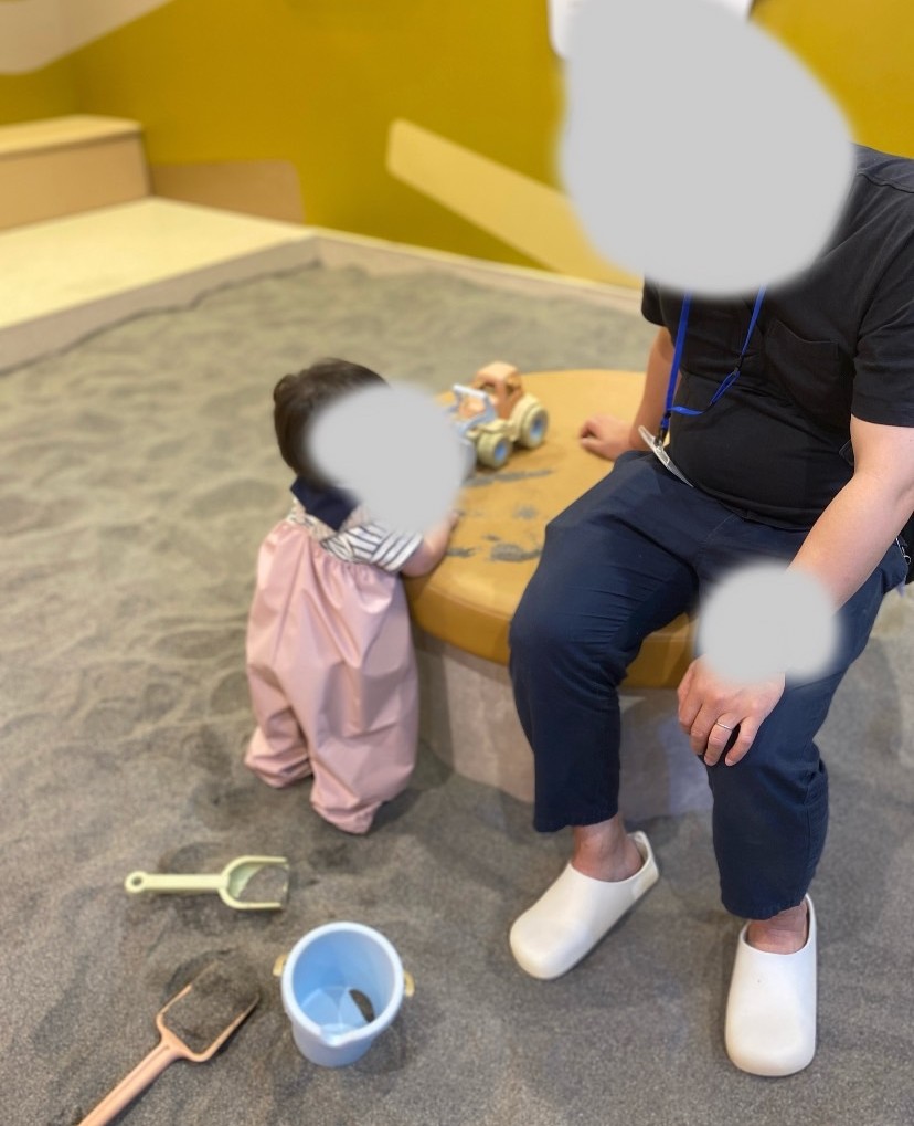 屋内の砂場で砂場着を着た1歳児がスタッフと遊ぶ様子。バケツやスコップも見える