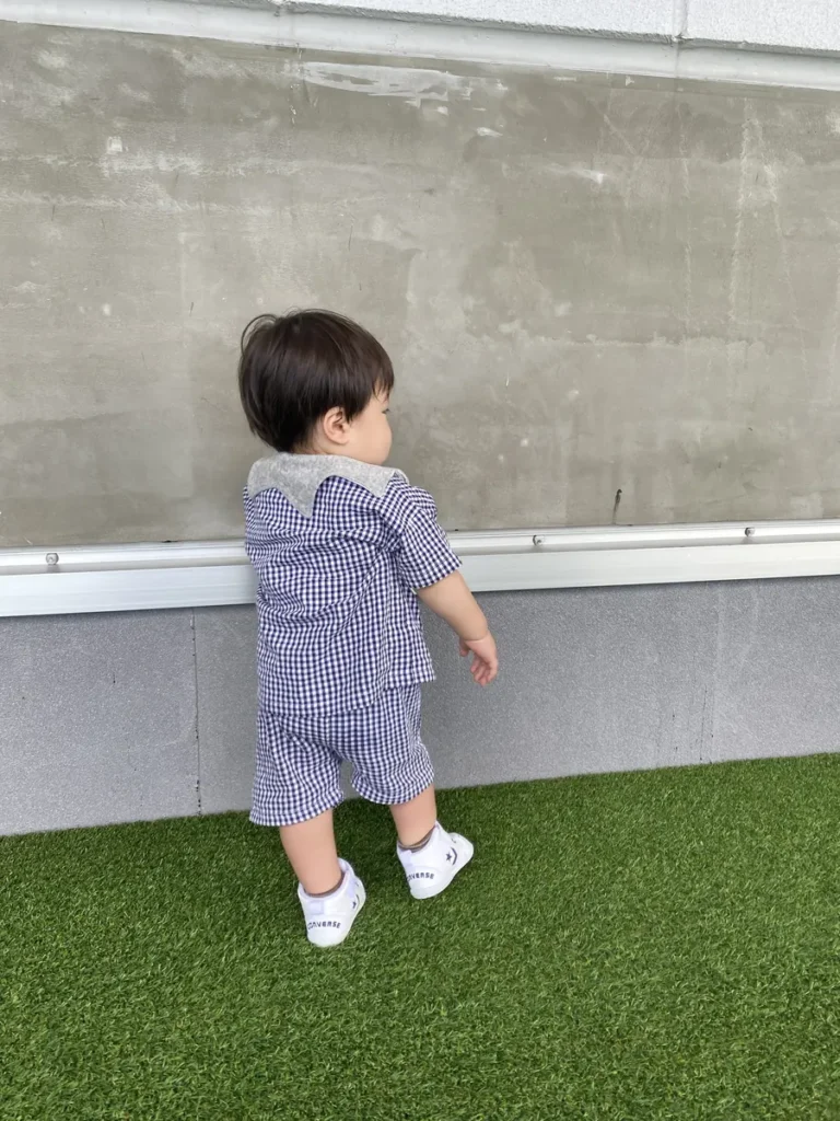 芝生の上で立つ1歳児の後ろ姿(チェック柄コーデと白スニーカー)