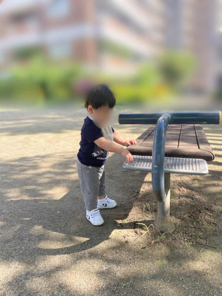 公園のベンチ横で立ち上がり、手をついて夢中で遊ぶ一歳児の様子を切り取った写真。